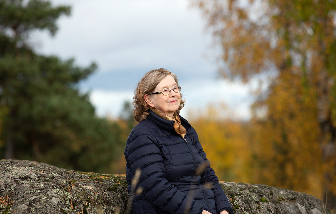 Osteoporoosia sairastava Helena Paulasto istuu kalliolla, hymyilee ja katselee kaukaisuuteen. Sää on syksyinen, 