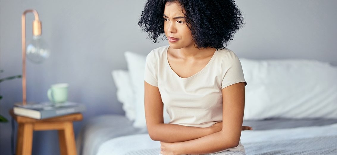 Kuvassa mustahiuksinen nainen istuu sängyllä ja pitelee vatsaansa molemmilla käsivarsilla. Ilme on jokseenkin tuskainen.